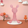 A pink silicone cartoon rabbit teether standing on a white base against a light pink background with clouds.