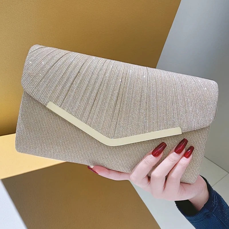 A hand holds a beige pleated glitter envelope clutch with a gold trim against a light background.