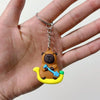 A small, cute brown bear charm with a blue shovel is attached to a silver keychain ring, held in a person's hand.