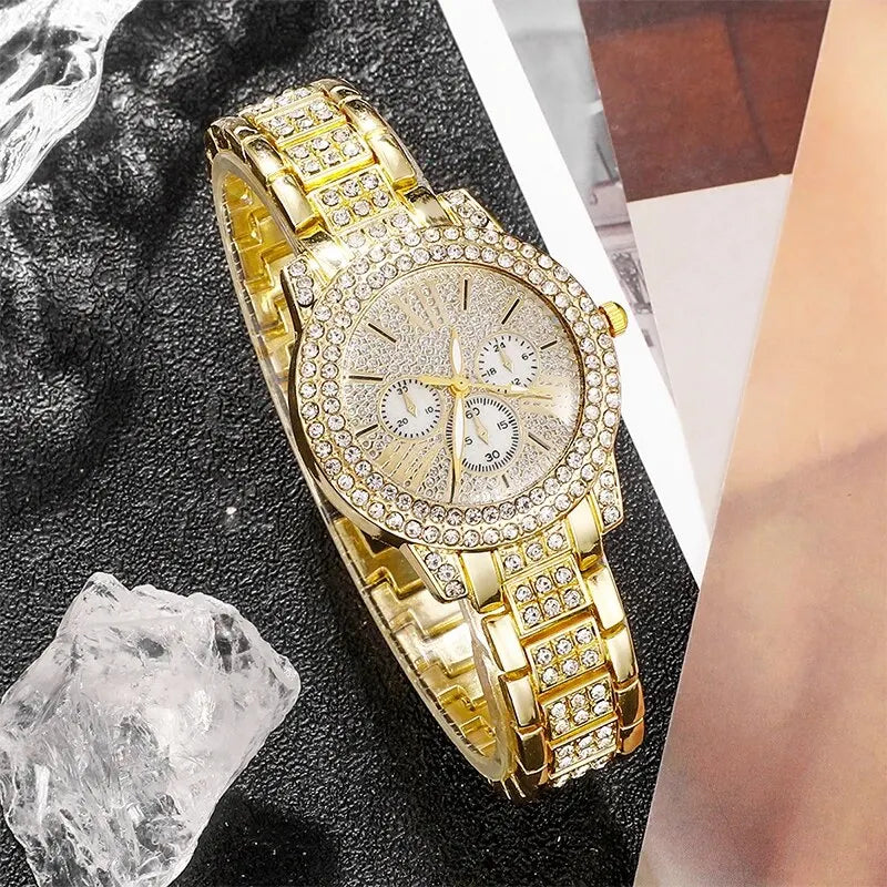 A close-up of a gold rhinestone watch and jewelry set resting on a dark surface next to a piece of ice.