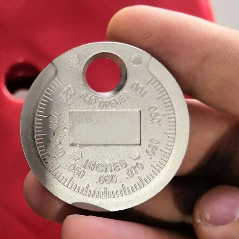 A hand holds a coin-type spark plug gap gauge showing measurements in inches and millimeters.