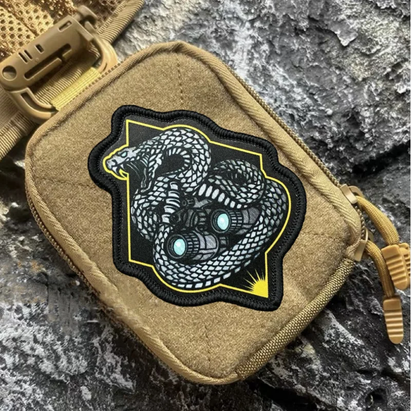 A tan tactical snake morale patch with a black and white design is shown attached to a beige hook-and-loop backing.