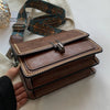 A brown leather mini cross-body bag with a patterned strap is shown next to a hand.