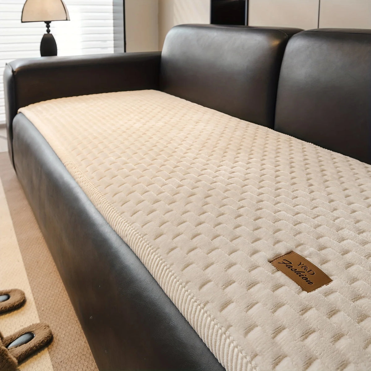 A dark leather sofa features a quilted beige plush cover with a non-slip backing.