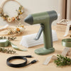 A green cordless hot glue gun with a white nozzle and cord lying on a wooden surface surrounded by natural greenery.