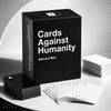 A black box of Cards Against Humanity Absurd Box party game with several cards laid out on a white surface.