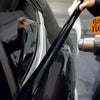 A hand is applying a glossy black vinyl wrap strip to the trim of a car.