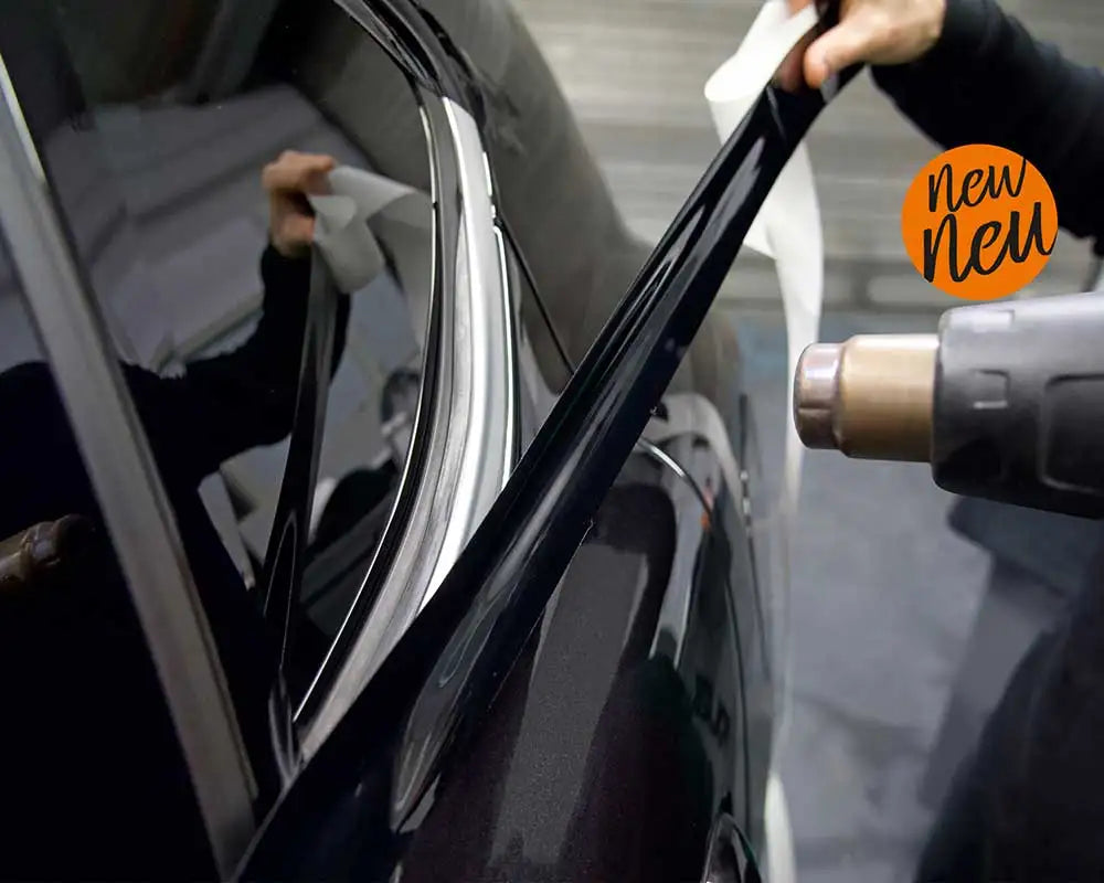 A hand is applying a glossy black vinyl wrap strip to the trim of a car.