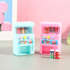 A pink interactive vending machine toy with clear compartments for play coins and drinks sits on a gray surface.