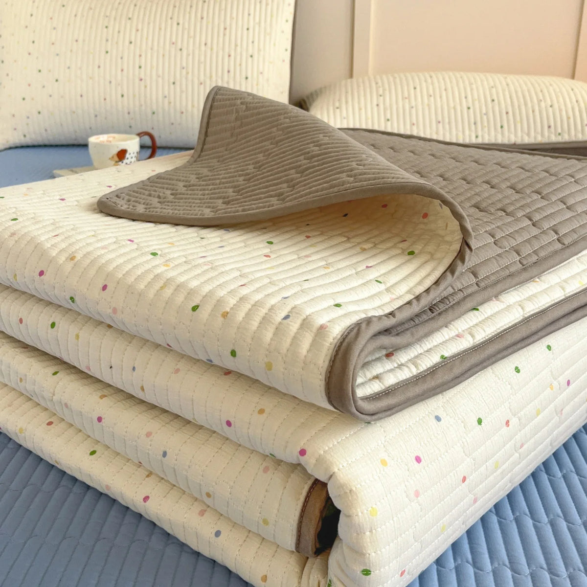 A stack of floral quilted bedspreads in light cream and gray tones resting on a blue surface.