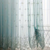 Light blue sheer curtain with silver floral embroidery hanging in a window.