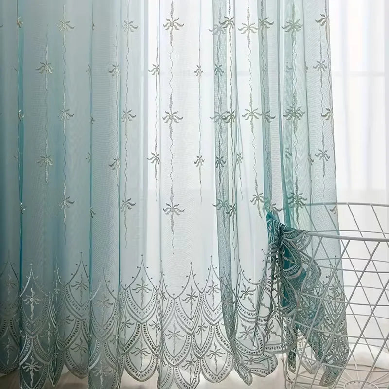 Light blue sheer curtain with silver floral embroidery hanging in a window.