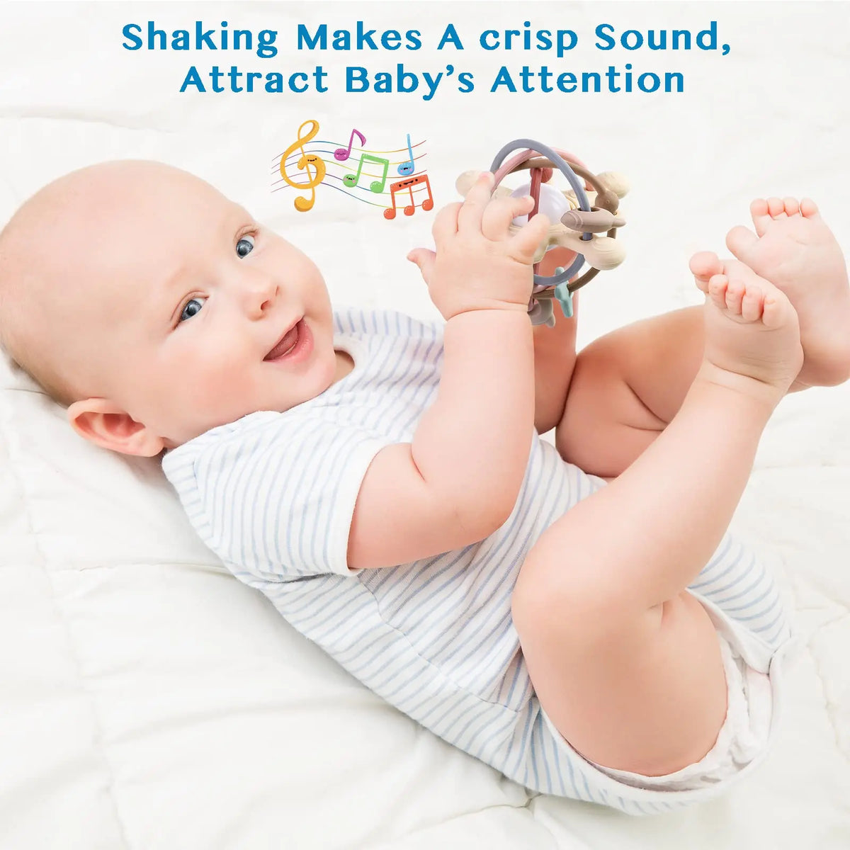 A smiling baby is holding a silicone sensory rattle ball while lying on a white surface.