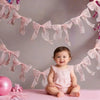A baby sitting on a pink surface surrounded by a garland of sheer pink ribbons and bows.