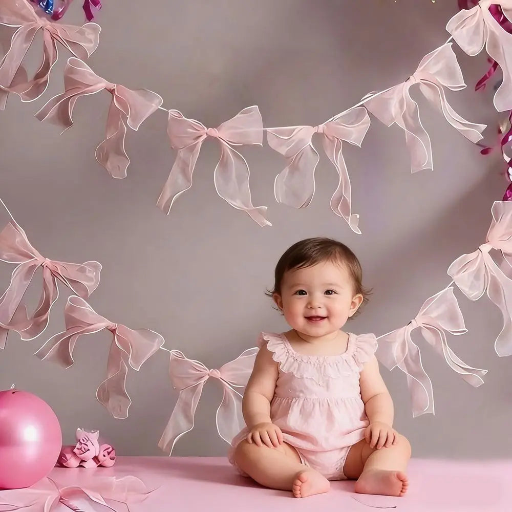 A baby sitting on a pink surface surrounded by a garland of sheer pink ribbons and bows.