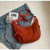 A burnt orange lightweight nylon crossbody bag with an adjustable strap resting on a denim shirt and a glass.