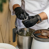 A person wearing black nitrile gloves is handling a metal container with a spoon in a bright kitchen setting.