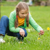 A young girl is kneeling in a green field, using an ergonomic stainless steel weeder set to pull small weeds.