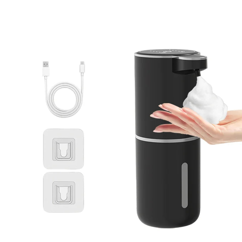 A hand is dispensing soap from a black touchless automatic soap dispenser, with charging cable and replacement parts shown ne
