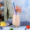 A hand holds a pink and blue stainless steel drinking straw next to a glass of iced beverage.