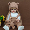 A lifelike silicone baby doll wearing a brown knitted bear hat and overalls with white shoes.