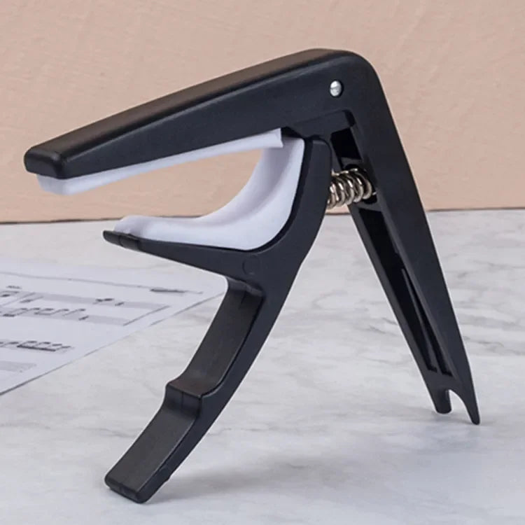 A black adjustable guitar capo with a built-in bridge pin puller shown on a white surface.