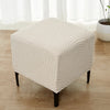 A beige geometric jacquard stretch ottoman cover placed on a wooden floor near a window.