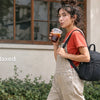 A woman wearing a red shirt and overalls carries a black mini backpack while holding a drink outdoors.