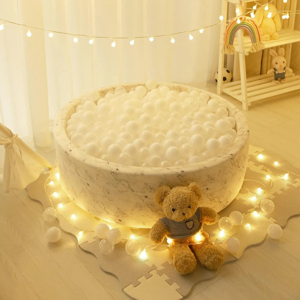 A large white ball pit filled with white balls is illuminated by string lights and features a teddy bear toy.
