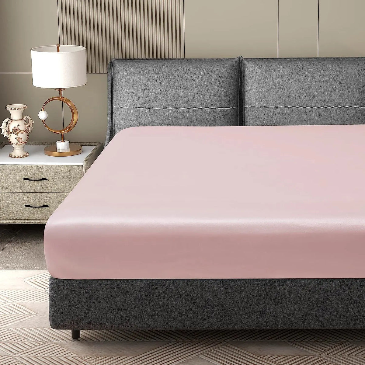A luxury satin fitted sheet is displayed on a bed with gray bedding and a bedside lamp.