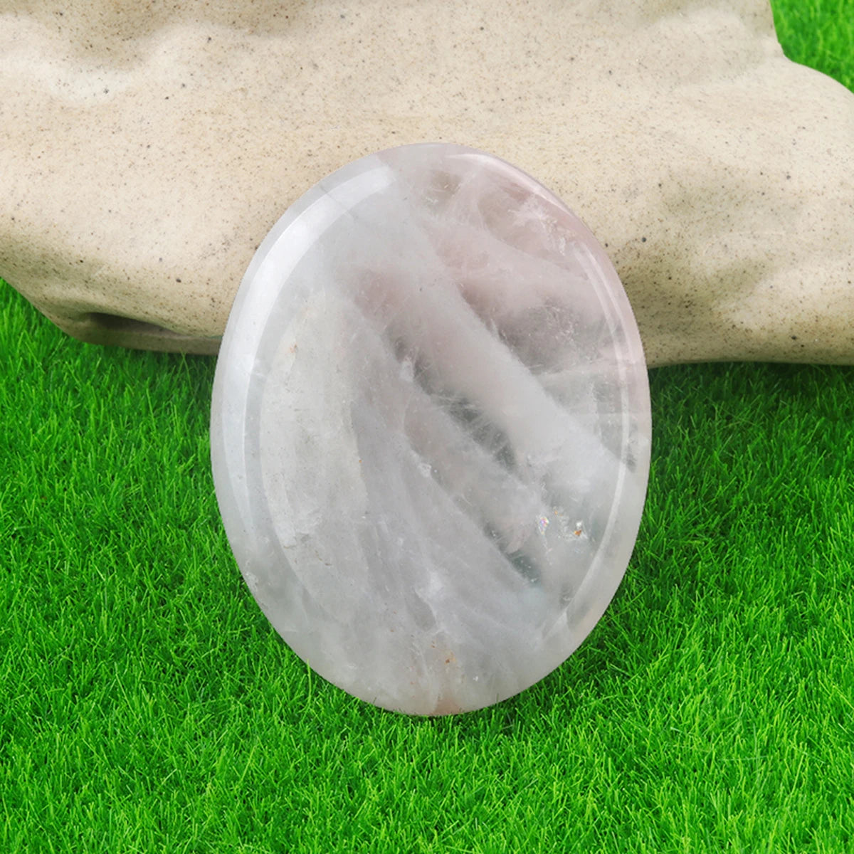 A smooth, oval, translucent white crystal stone resting on green grass next to a hand.