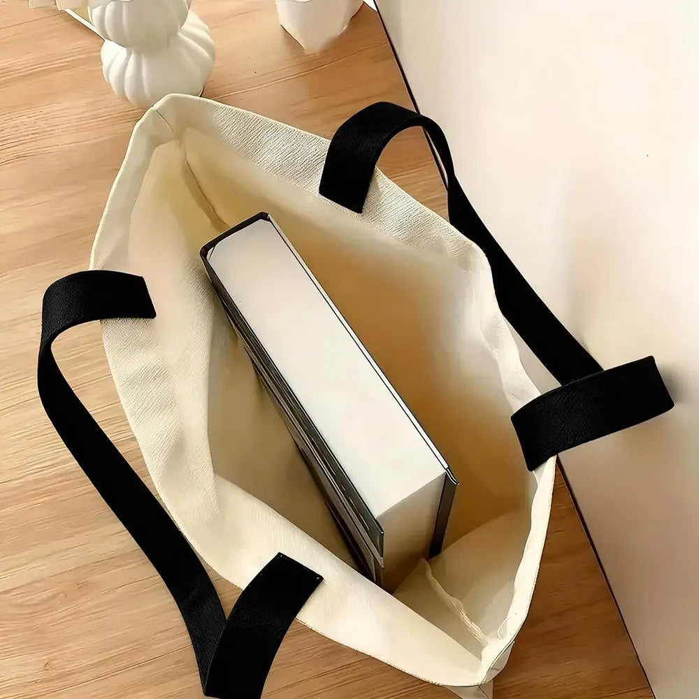 A cream canvas tote bag with black straps holds a white rectangular item on a wooden surface.