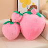 A pink strawberry plush throw pillow with green leaves and a brown stem, displayed on a light-colored surface.