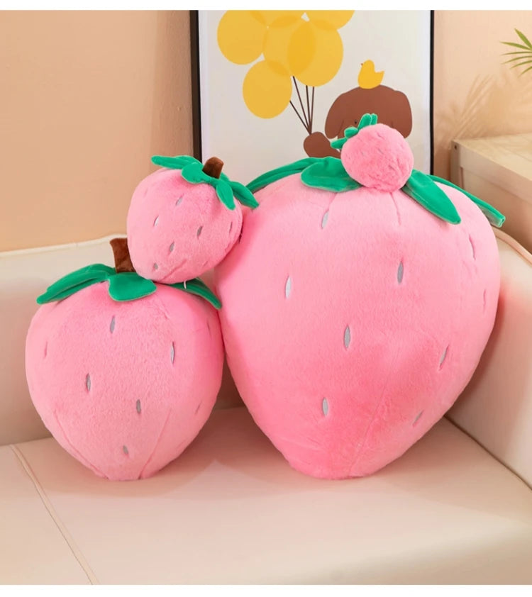 A pink strawberry plush throw pillow with green leaves and a brown stem, displayed on a light-colored surface.