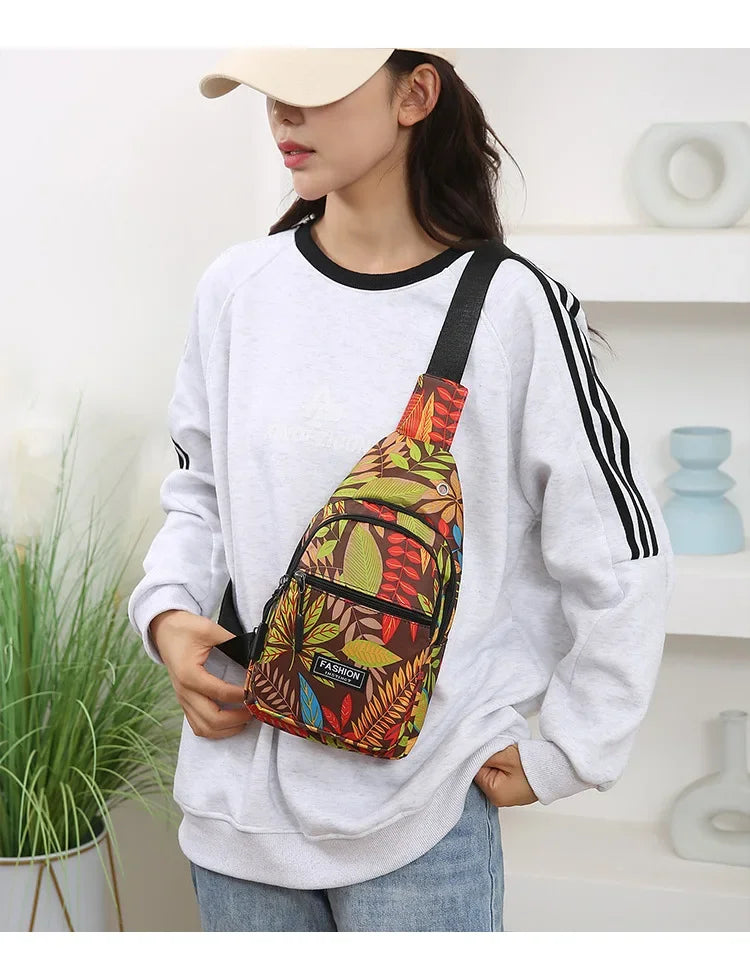 A woman wearing a white sweatshirt holds a colorful leaf print sling crossbody bag with multiple pockets.