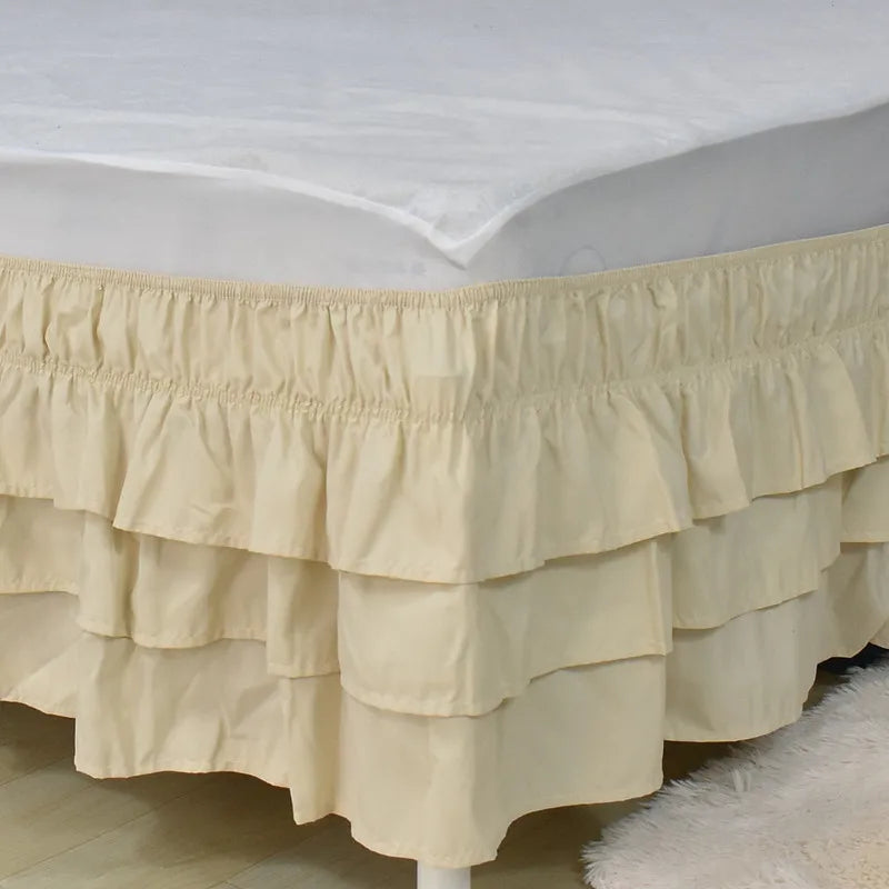A beige adjustable elastic bed skirt with ruffled tiers is shown covering the edge of a mattress.