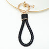 A gold-toned key fob cover featuring a black braided leather strap attached to a thin gold chain.