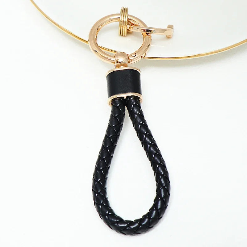 A gold-toned key fob cover featuring a black braided leather strap attached to a thin gold chain.