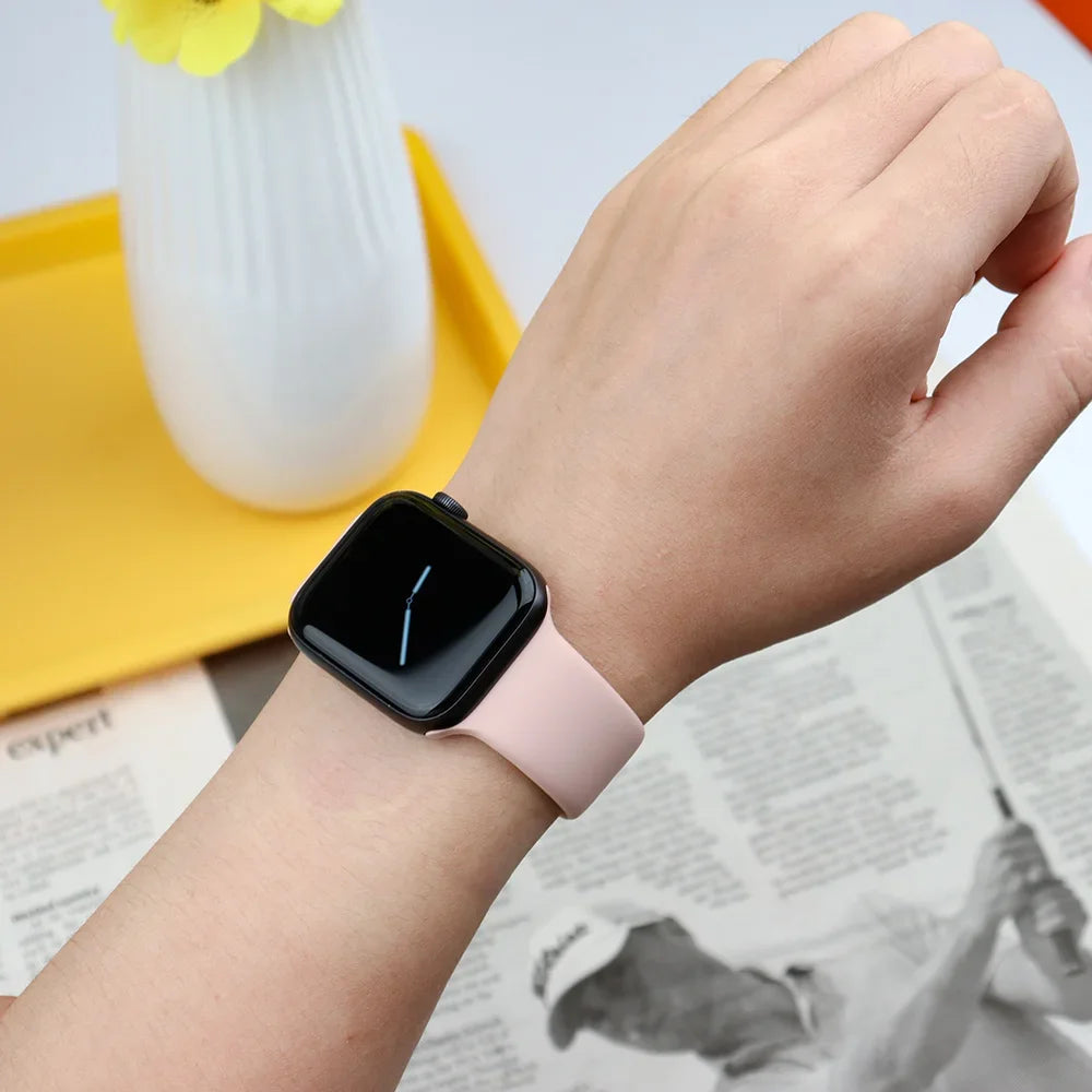 A hand wearing a pink silicone sport band on an Apple Watch, next to a yellow plate and some printed paper.