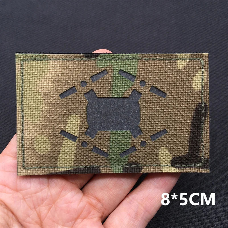 A rectangular camouflage tactical patch with a national flag design and hook-and-loop backing is held in a hand.