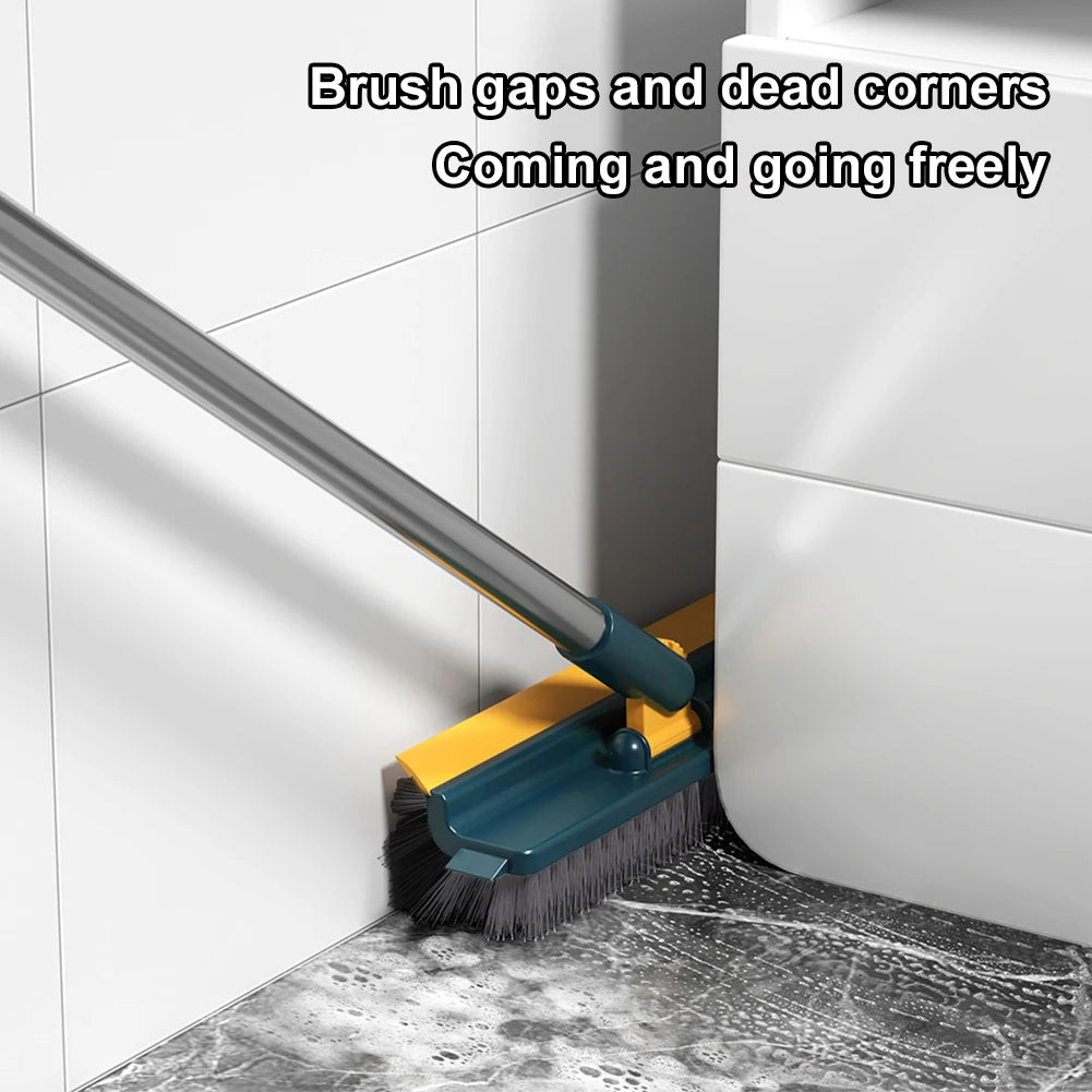 A yellow floor scrub brush with a long handle is cleaning a corner gap in a white cabinet and tile surface.