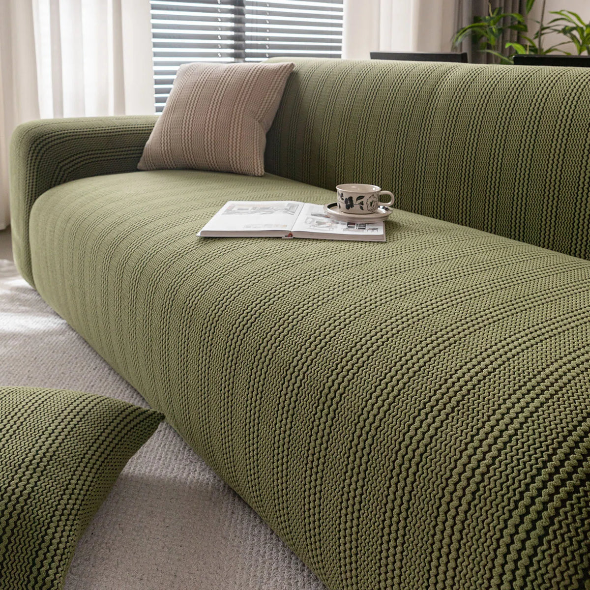 A textured olive green quilted stretch sofa cover with a light beige throw pillow and an open book on the surface.
