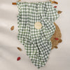 A muslin baby swaddle blanket in a green and white checkered pattern is displayed on a light background with dried leaves.