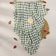 A muslin baby swaddle blanket in a green and white checkered pattern is displayed on a light background with dried leaves.