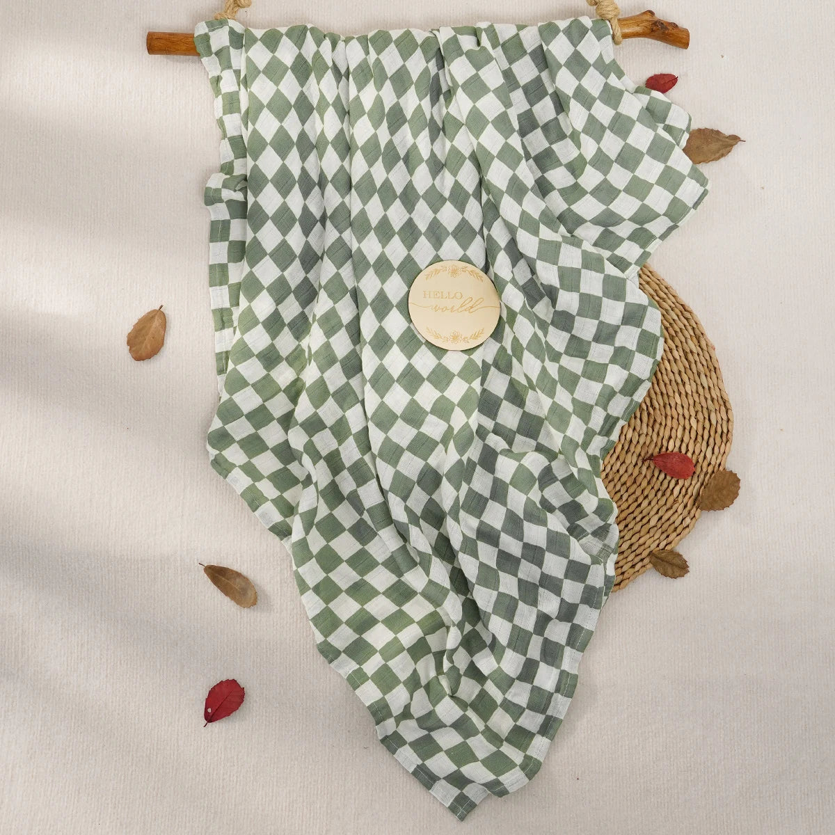 A muslin baby swaddle blanket in a green and white checkered pattern is displayed on a light background with dried leaves.