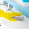 A yellow glove is holding a gray pumice stone near a white sink and faucet.
