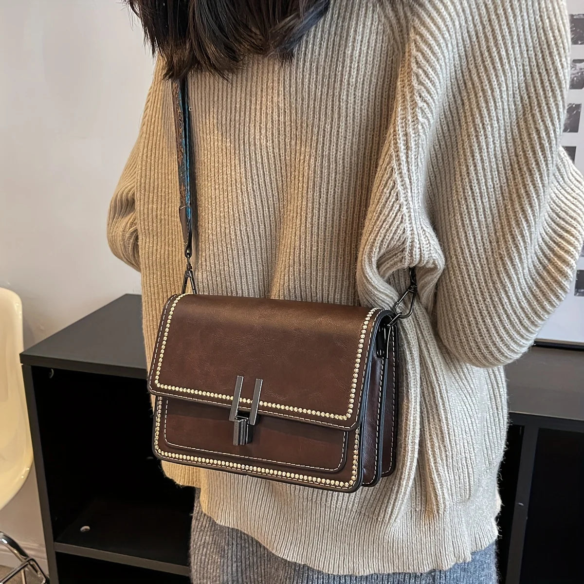A brown leather mini cross-body bag with a gold chain is shown against a light beige knit sweater.
