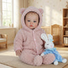 A soft pink reborn baby doll with a hood and rabbit ears is sitting on a light rug.