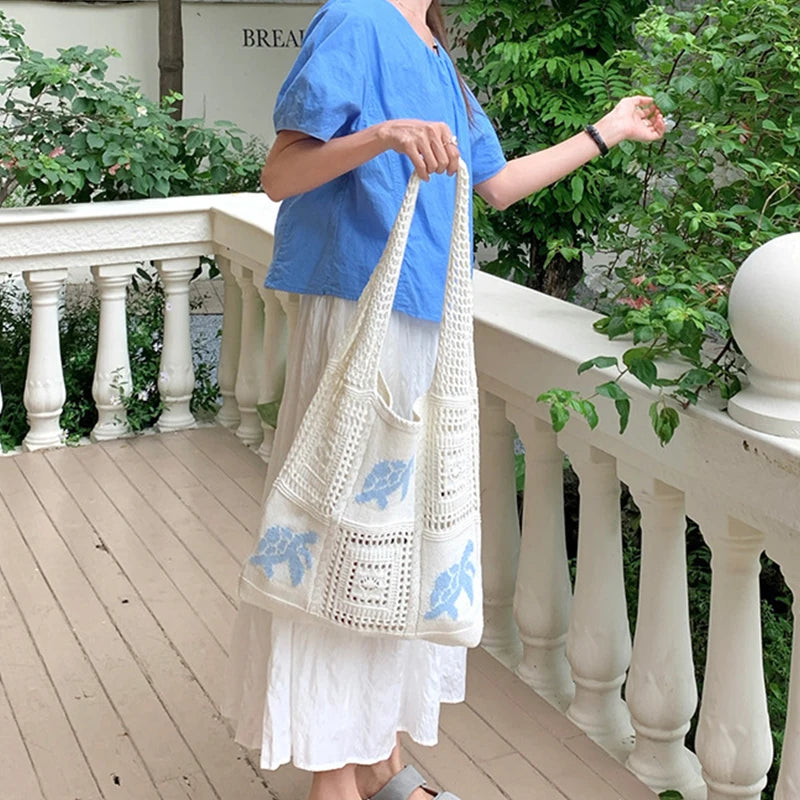 A person holds a white handwoven knit tote bag with blue embroidery on a porch railing.