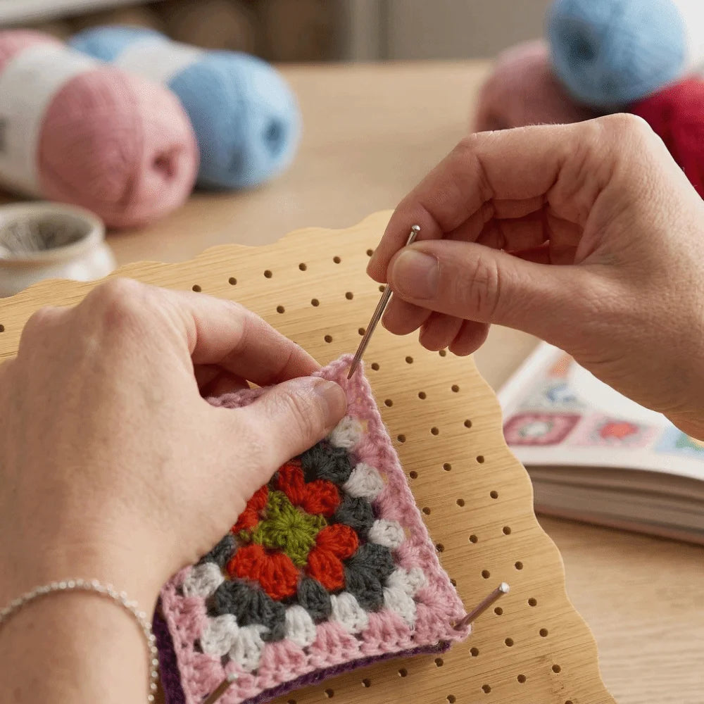 Hände verwenden eine Häkelnadel, um ein rosa-lila gestricktes Stück auf einem Holzblock zu bearbeiten.